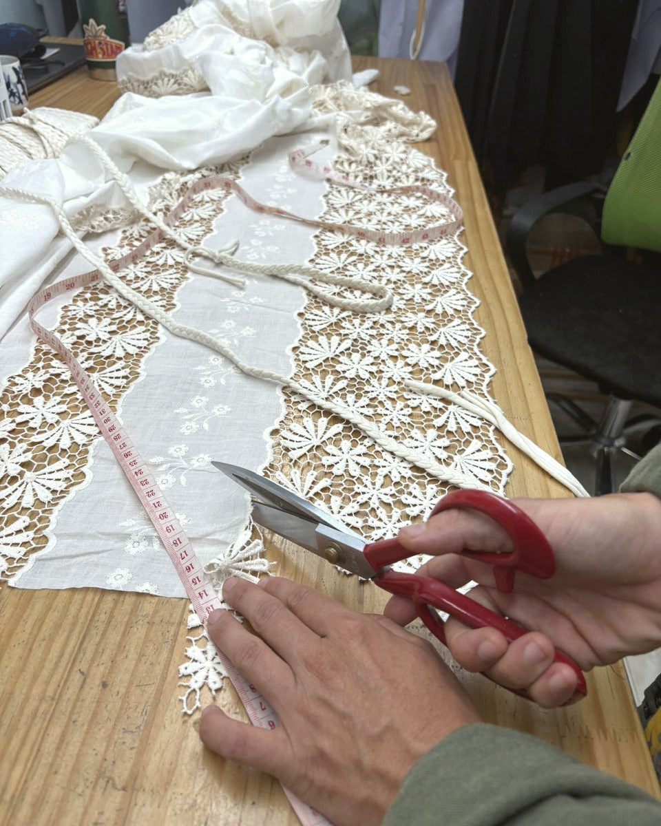 Hands cutting lace fabric with scissors along a measuring tape on a workshop table