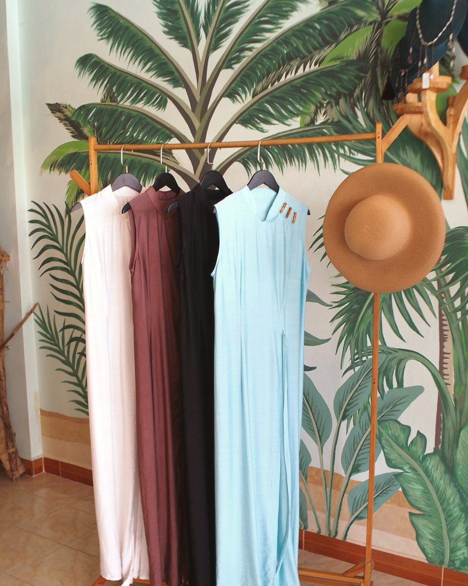 Four sleeveless dresses and a straw hat on a wooden rack in front of the palm-leaf mural in the Best Plan No Plan atelier, Koh Phangan