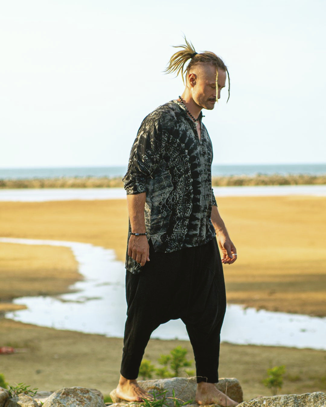 Man in a tie-dye shirt and dark harem pants walking over rocks above a shallow tidal flat at sunset