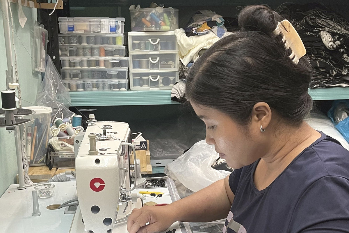 Seamstress sewing at an industrial machine in the atelier, shelves of thread and fabric behind her